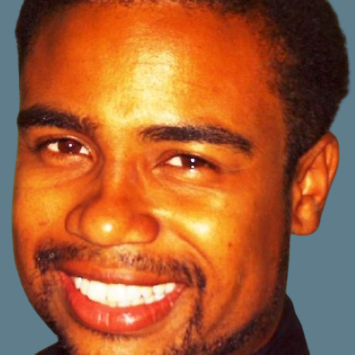 Foto de perfil de homem sorrindo com pele parda e óculos de sol com um fundo na cor azul.